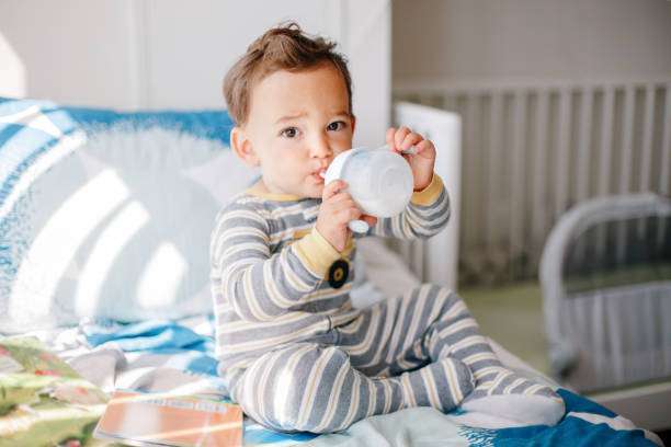 The baby is sitting on the bed with the bottle warmer on.