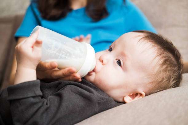Mother, feeding her baby boy from bottle warmer comfortably sitting on the couch at home