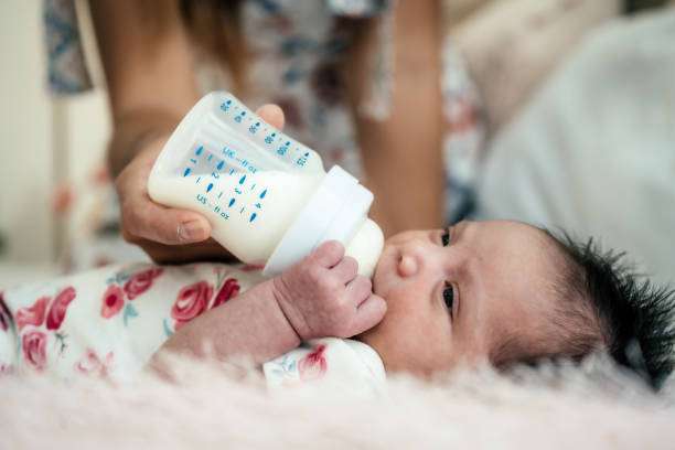 Baby girl drinking milk from a bottle warmer with mother sitting by on floor.