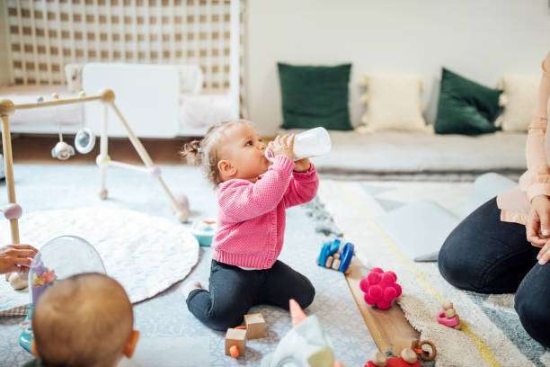 Baby girl drinking milk from a bottle warmer with mother sitting by on floor.